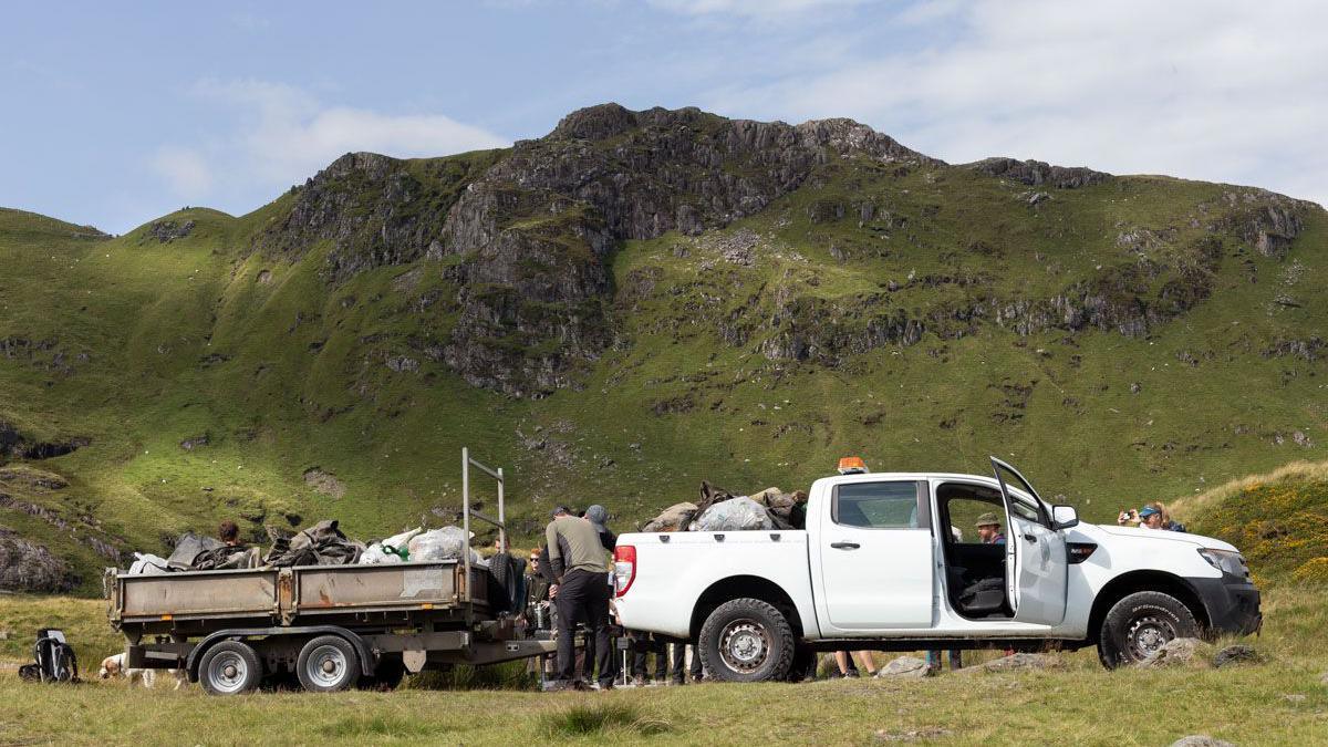 A white van with a trailer, parked next to a green mountain range. There are people surrounding the trailer which is piled with rubbish bags,