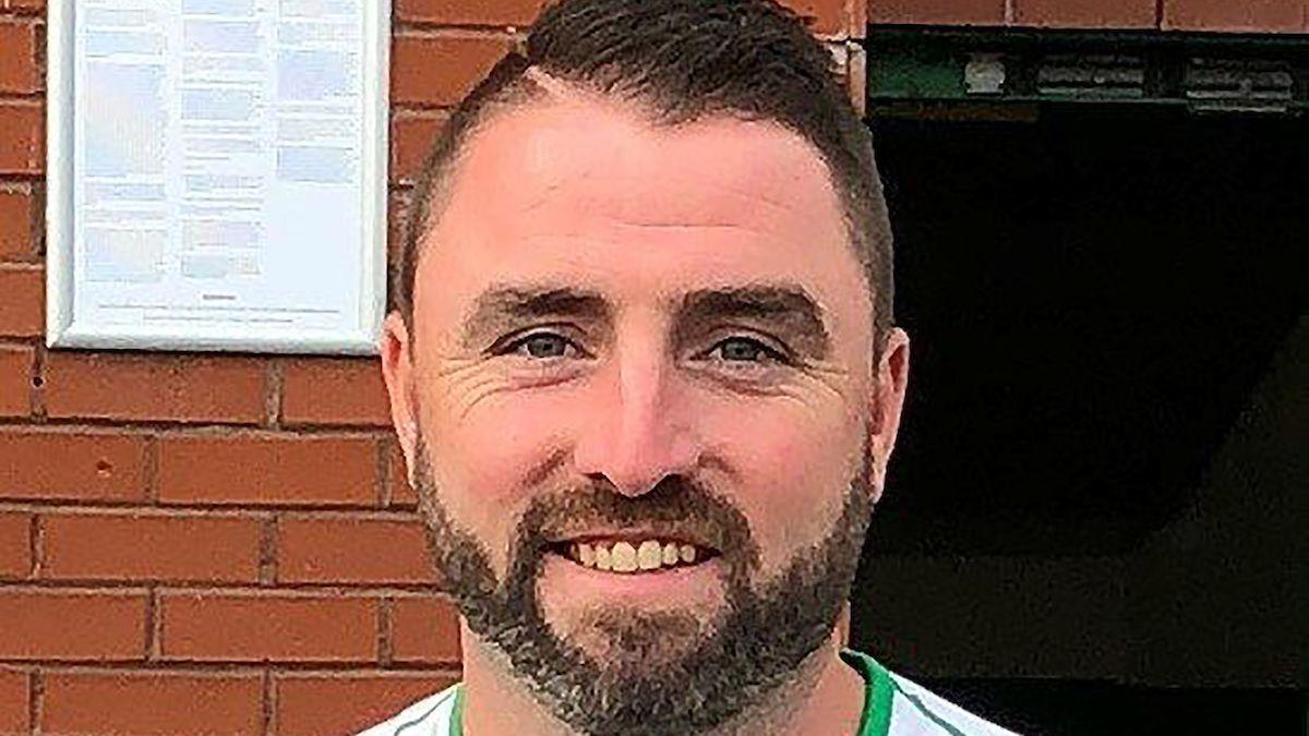 Jim is smiling at the camera in front of a red brick wall with a white sign on it with words that are too small to make out. 
He has brown hair brushed to the side and a brown beard.
He has blue eyes and is wearing a white sports top that is cut off in the crop. 