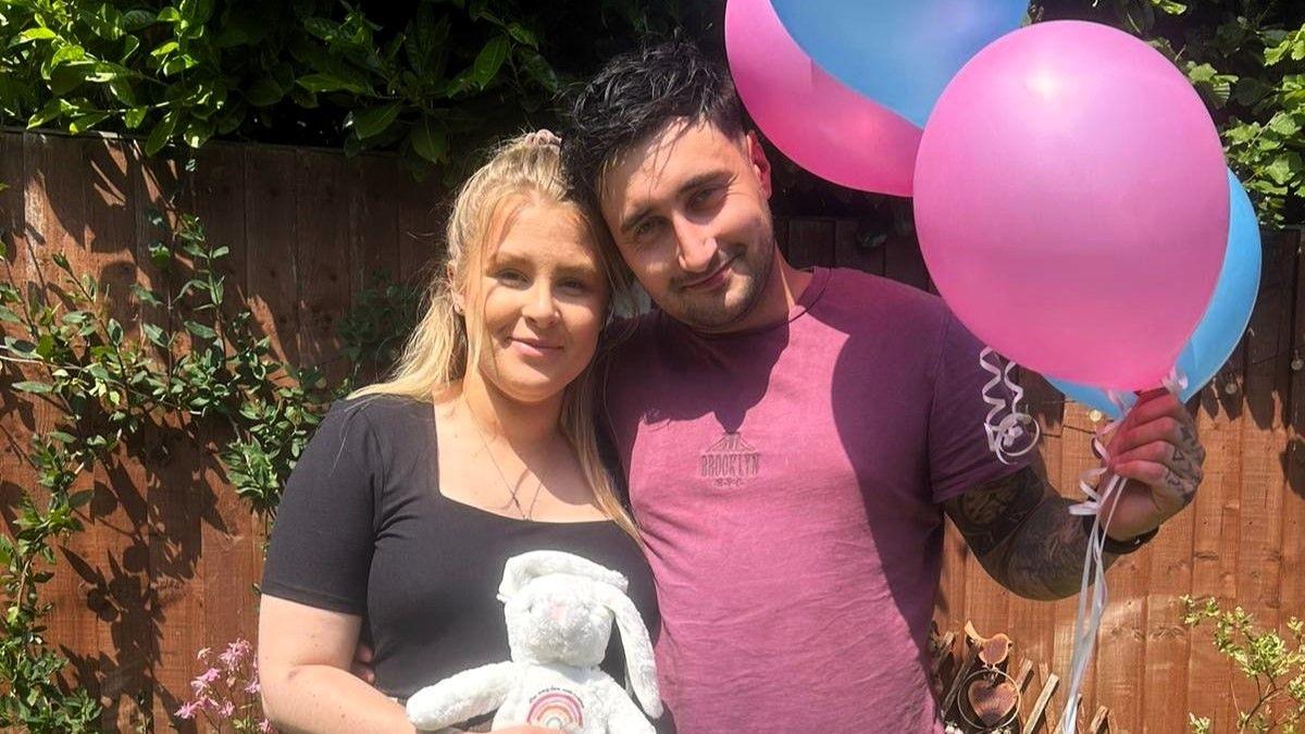 Oliver Dent wearing a plum coloured t-shirt and holding pink and blue balloons. He is stood next to Caitlin who/s wearing a black t-shirt and holding a toy rabbit.