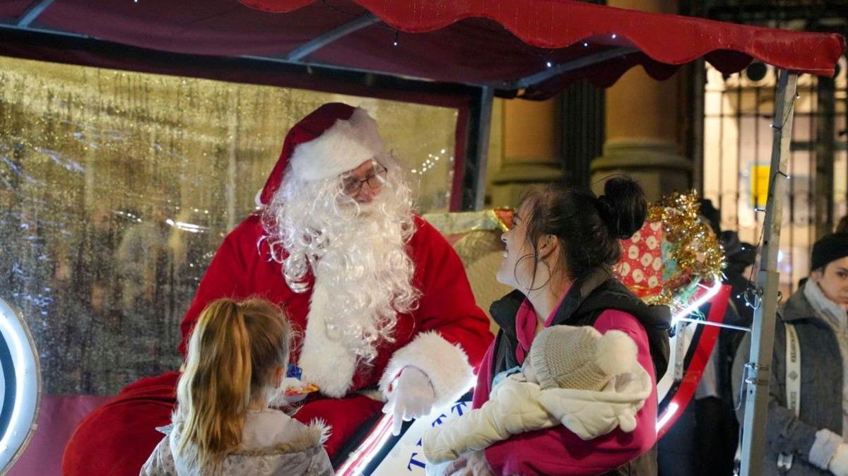 A man wearing a red outfit and white beard, Father Christmas, is sitting in a sleigh with a present behind him. He is talking to a woman, who is holding a baby, and a girl is standing next to them.