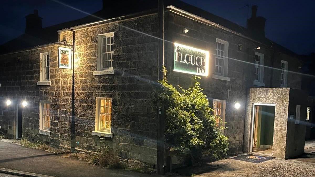 The image shows a square Georgian building at night with the name, the Plough inn lit up 