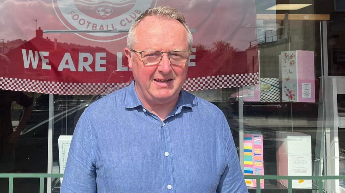 A man in a blue shirt stands in front of a shop window. In the window is a Larne flag. The flag is mostly red, with the Larne crest in the middle and ‘We are Larne’ written in white letters below the crest. The Larne crest is a white circle with a red middle. In the middle is a white sail boat.

