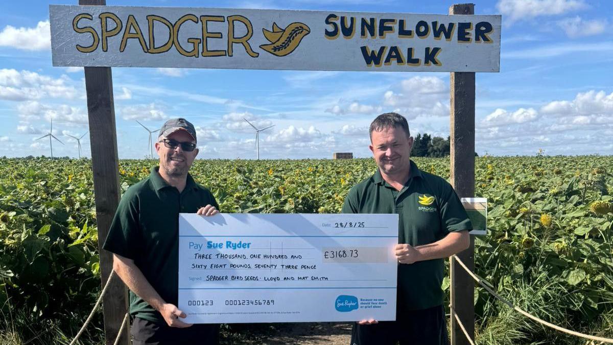 Two men wearing green T-shirts are holding a big cheque while standing in a sunflower field. The man on the left has a cap and sunglasses on.