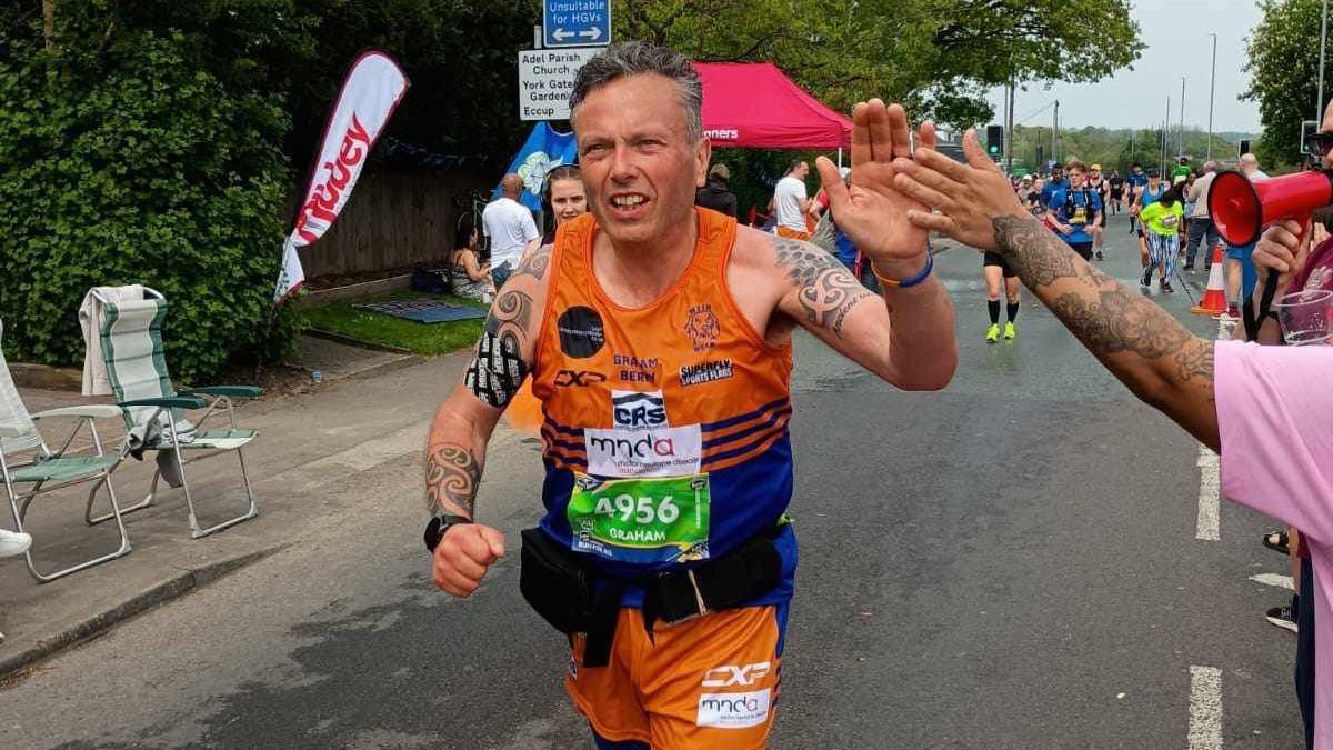 Graham Berry running the Leeds Rob Burrow Marathon. He is wearing blue and orange and there are other runners behind him. He is holding his hand up to greet a spectator with a high five.