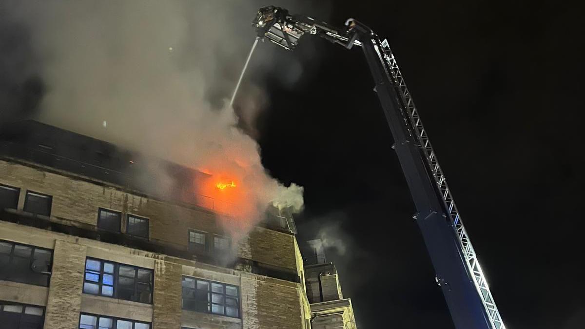 Fire crews use an extension ladder with hose as they spray the roof of an abandoned building. Fire and smoke can be seen billowing from the roof.