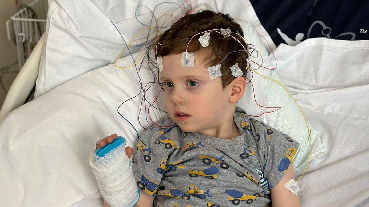Samuel lies in a hospital bed with electrodes attached to their head via colorful wires. The child has a bandaged hand and wears a gray shirt with yellow construction vehicles. Medical equipment surrounds the bed, including a monitor showing vital signs