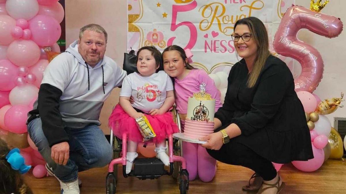 Paul McRae, Amber, Lalia and Leanne at Amber's fifth birthday - Leanne is holding a cake while there are pink and white balloons and banners behind the family, including one as a giant 5.  