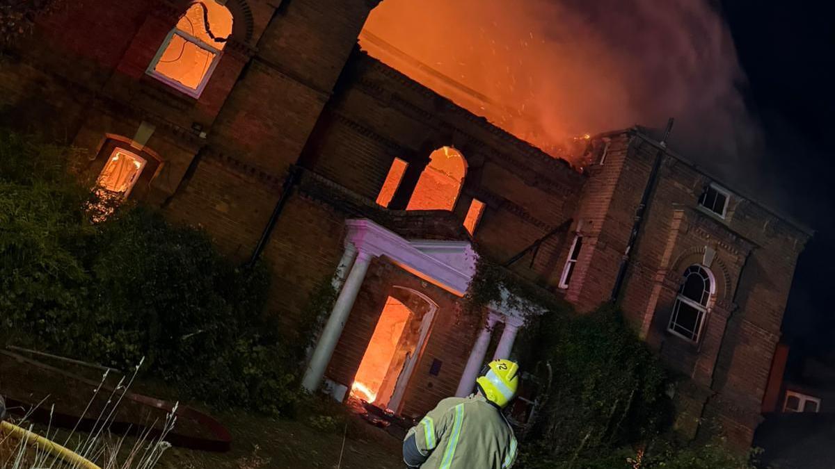 A firefighter standing in front of a large double fronted property. The property is shown gutted by a fire which is visible and illuminating the interior of the structure. The roof of the property is severely damaged and some of the windows are broken.