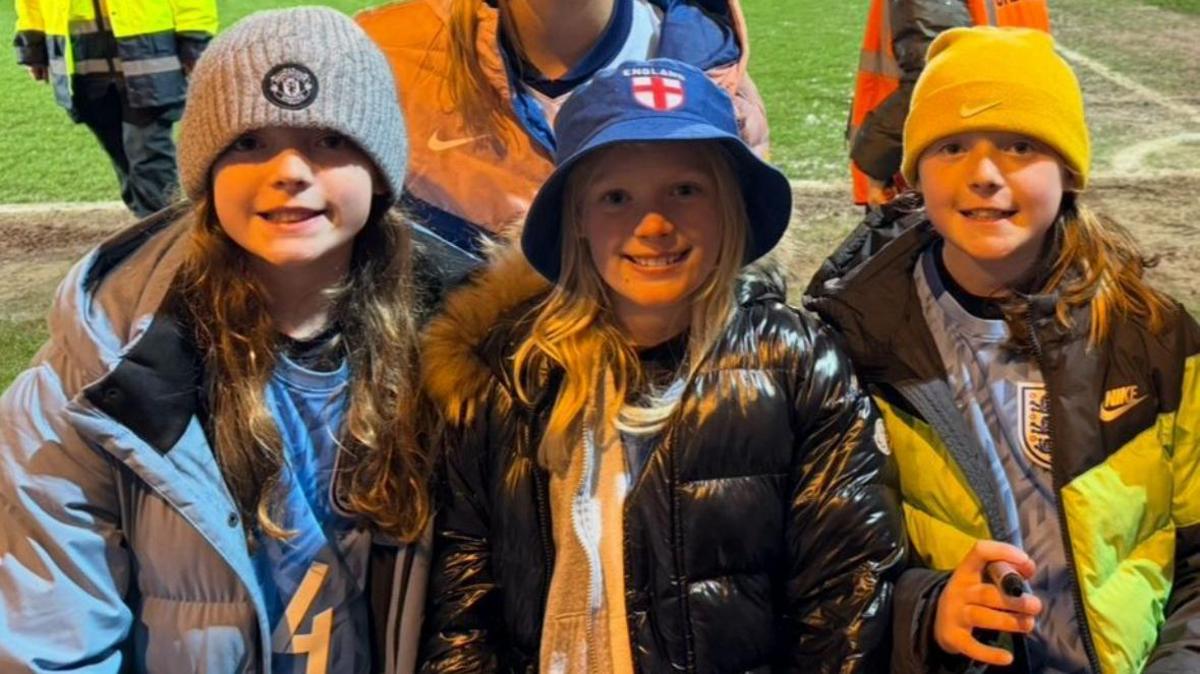 Three young girls are standing on the edge of a grass football pitch. They are wearing padded coats and hats. One has a Manchester United beanie on, another has an England bucket hat on, and the other has a Bike Beanie on.
