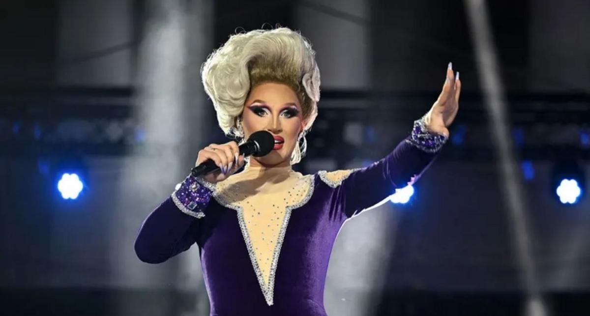 Mae perfformiwr mewn gwisg borffor ar lwyfan yn dal meicroffon ac yn chwifio at y dorf