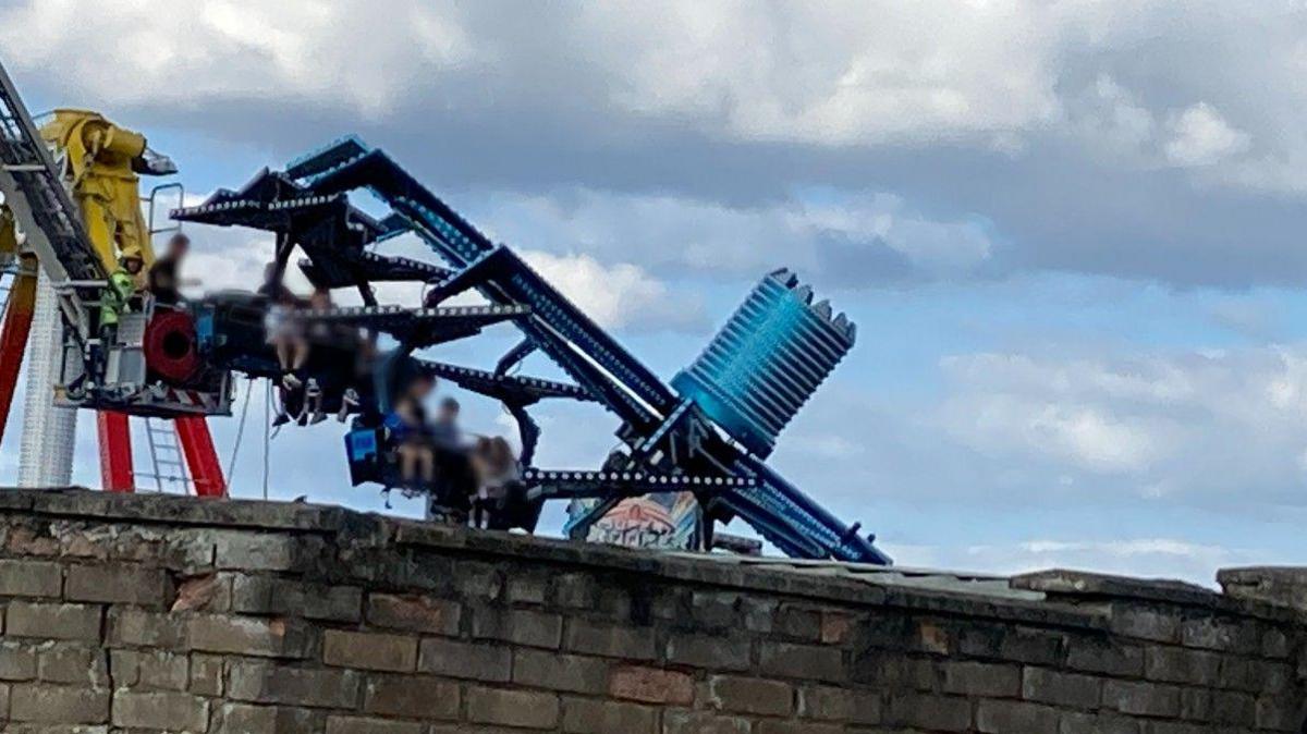 Adults and children suspended in mid-air on blue-coloured ride with wall in foreground