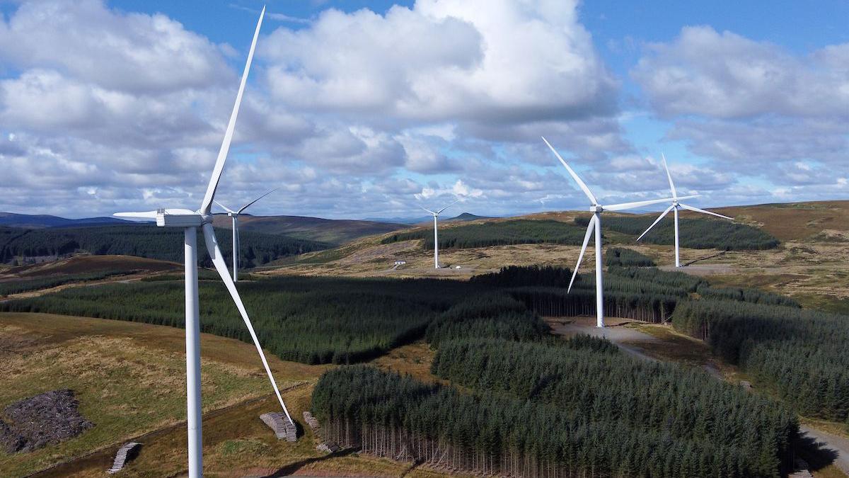 A set of wind turbines on a Borders hillside surrounded by stretches of forest and a cloudy blue sky