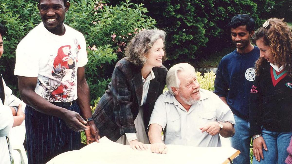 Lee Durrell and Gerald Durrell at a table, Lee Durrell is stood above her husband, who is sat down. They are talking to someone next to them, and are surrounded by other people