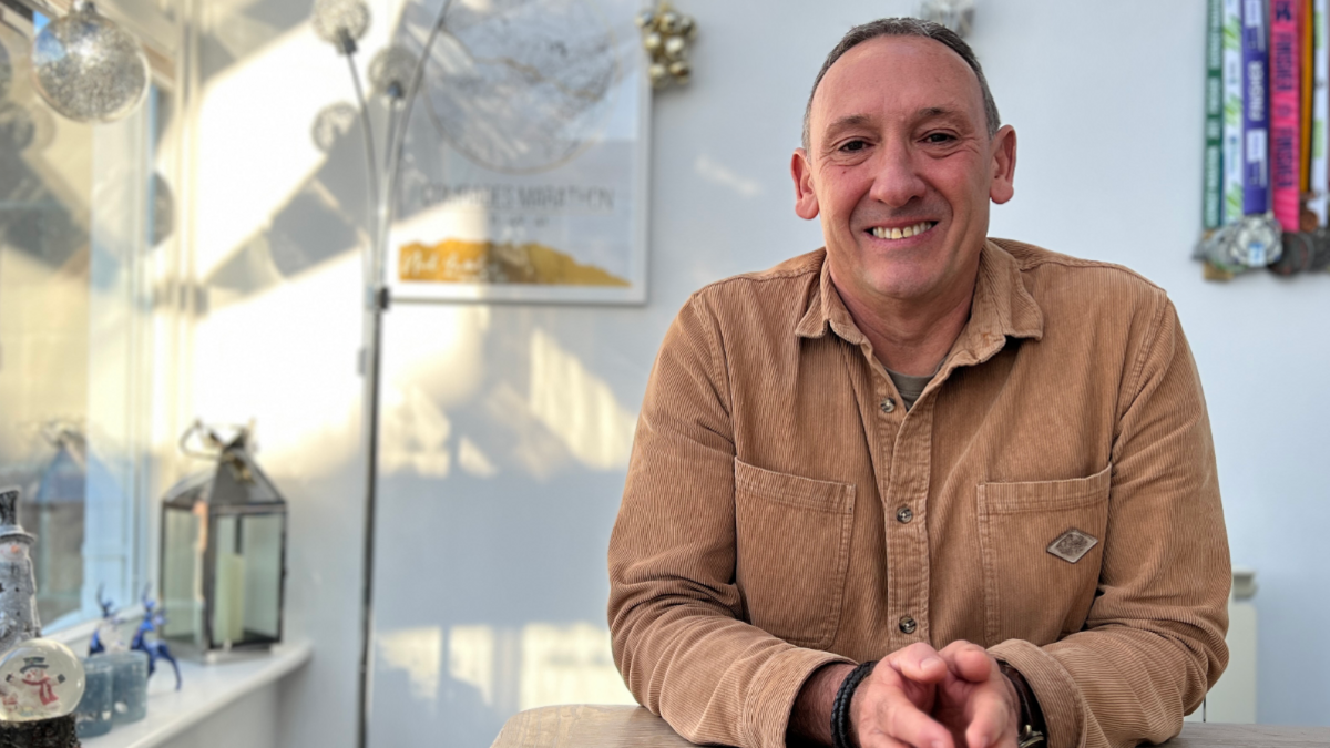 A smiling man with a brown shirt with a background of house lamp and some window lights.