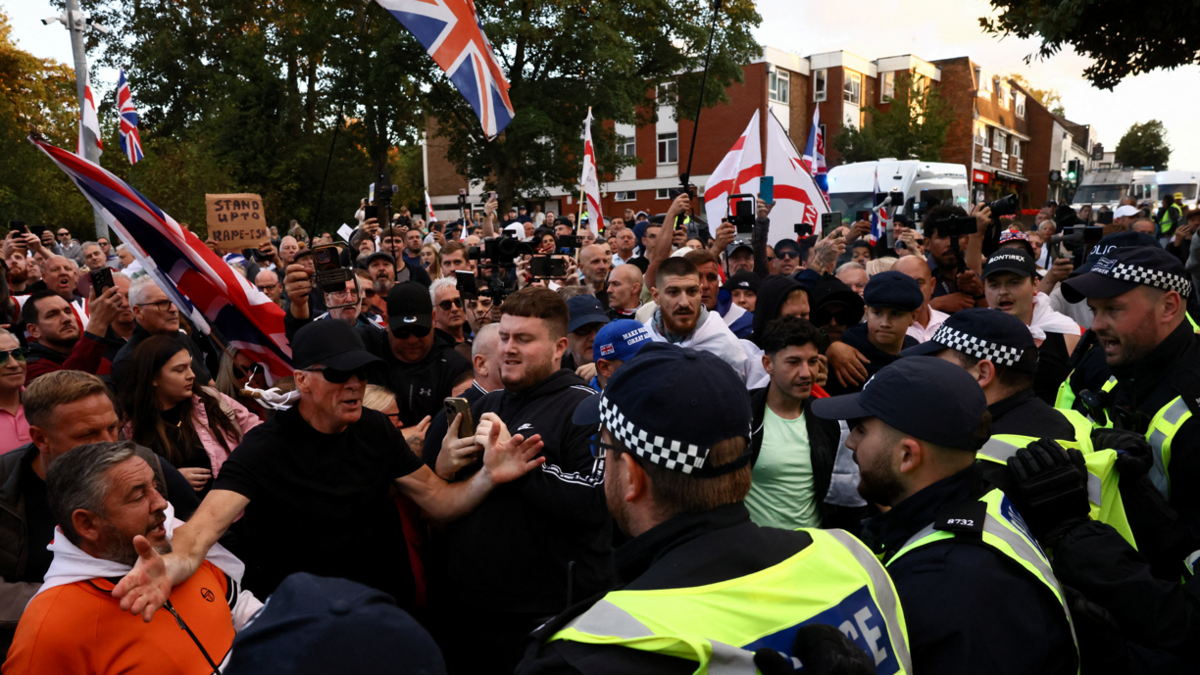 Large gathering of people some holding union jack and st georges cross flags some facing a line of police officers.