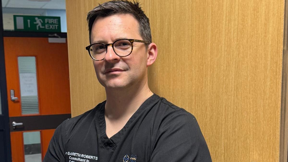 Gareth is stood against a door in the hospital, he has short dark hair and is wearing dark-rimmed glasses. He is wearing black hospital scrubs with his name and the NHS logo embroidered on.