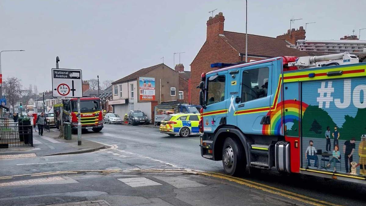 A busy road in central Hull with shops and residential houses on each side. A blue fire truck can be seen on the right, along with a red one further up the road. A police car is also parked in the middle of the road.