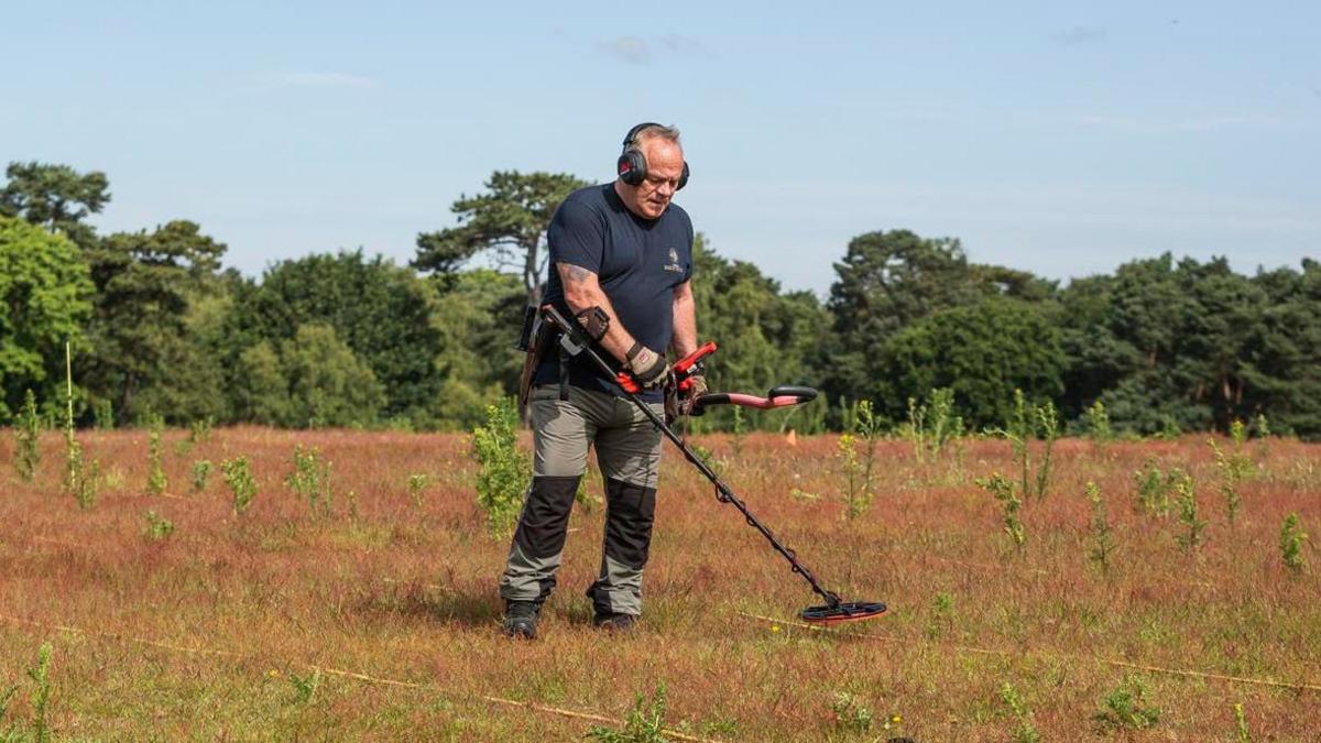 A Time Team member is pictured working on site at Sutton Hoo in a field. He is using a metal detector to scan the ground for any historical items. He is also wearing headphones over his ears. Trees can be seen in the background behind him.