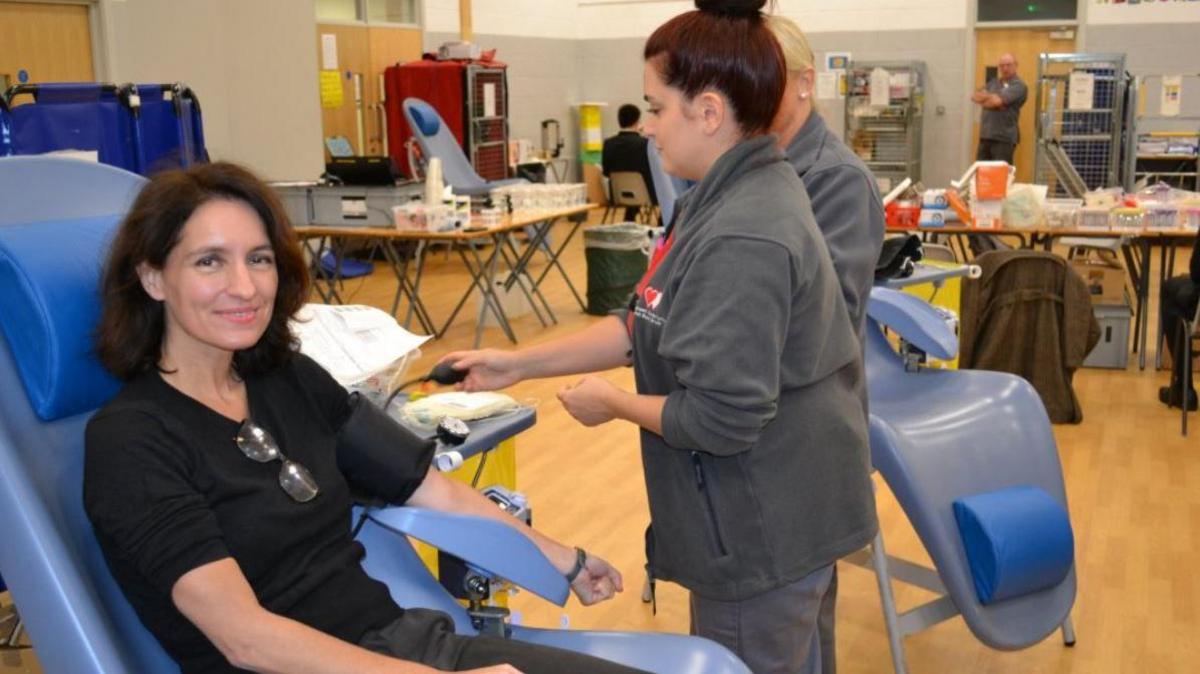 A woman sits on a chair in a school hall smiling at the camera. She has a blood pressure cuff on her arm which a Welsh Blood Service nurse is inflating. Tables of medical supplies are visible in the background.
