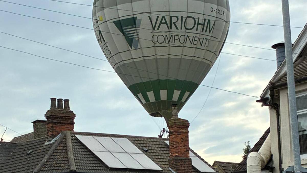 A hot air balloon hovering over roofs in Bedford. The balloon is green and grey and you can see solar panels, chimneys, poles and wires.