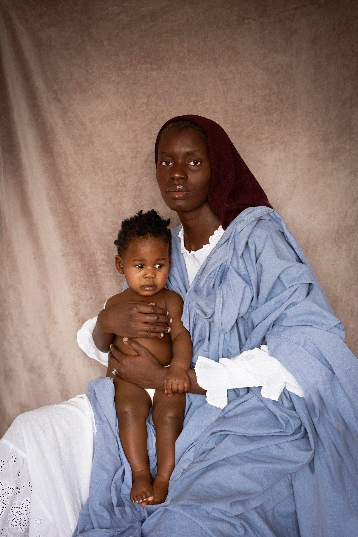 Dola Posh in a black headscarf and wearing a blue cloth over a white dress stares with an empty expression at the camera. She is holding her daughter in her hands.