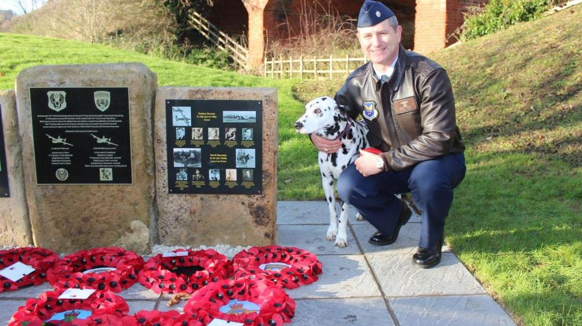 Lt Col Chris Nastal pictured at the memorial with a Dalmatian dog with wreaths in the foreground.

