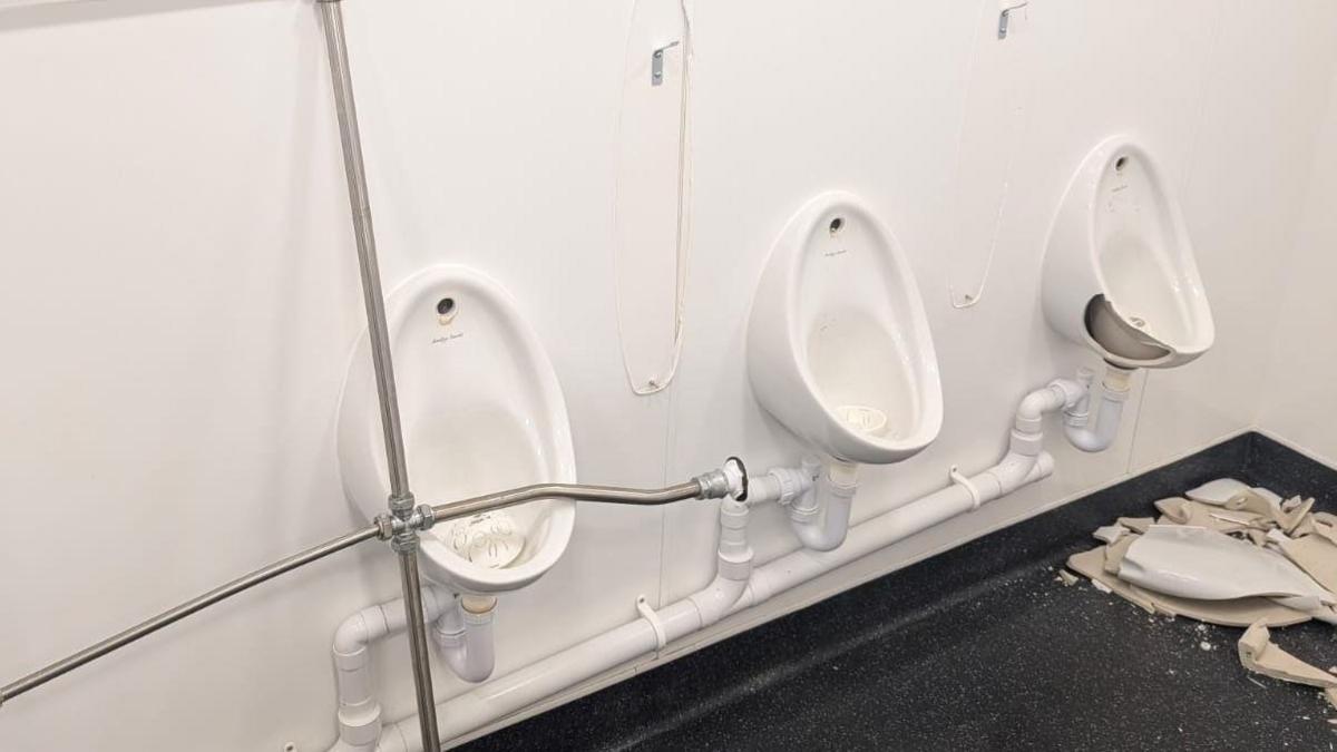 Three white urinals are on a white wall in a public toilet. The one at the far right is badly chipped and there is a pile of broken porcelain on the dark floor beneath it