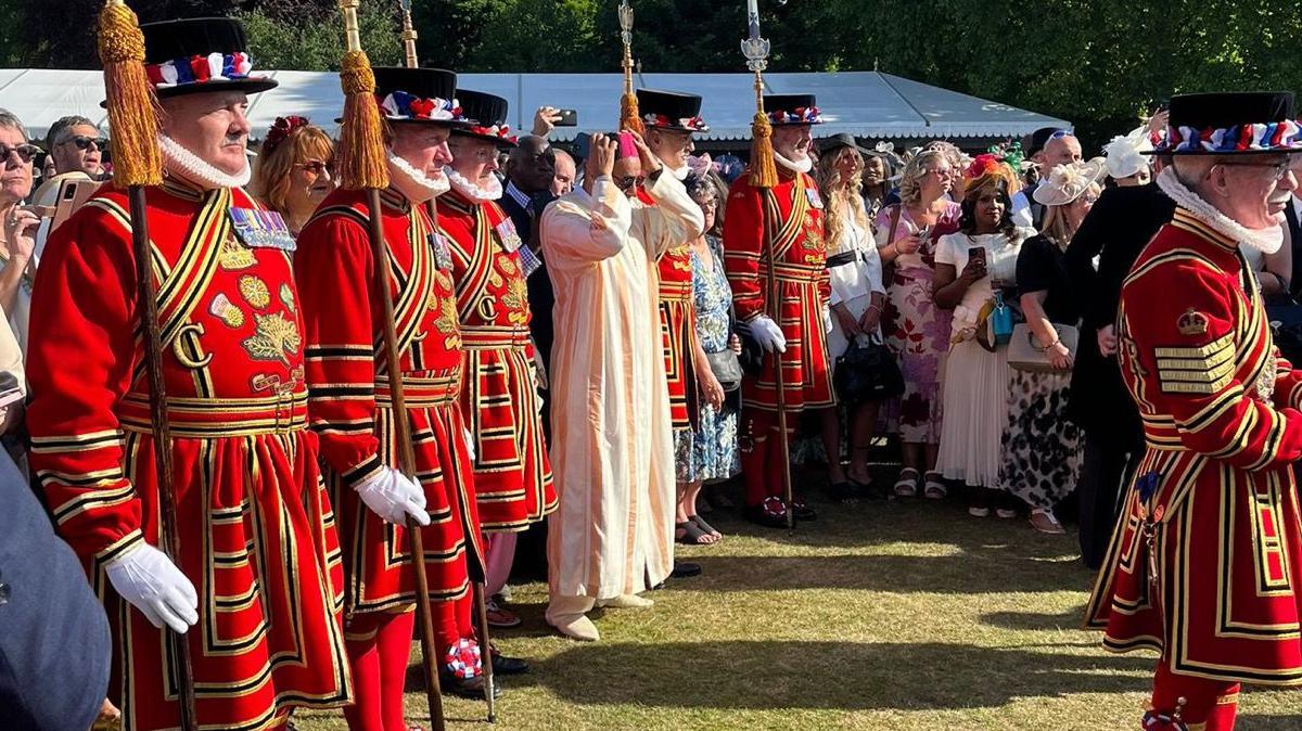 Six beefeaters are seen amongst a crowd of people all smartly dressed. They are in bright red and gold clothing, with staffs adorned with a gold tassel in their hands. They wear black hats, with red, white and blue flowers around the rim. A man with a pink hat and a cream dress is seen in the middle.