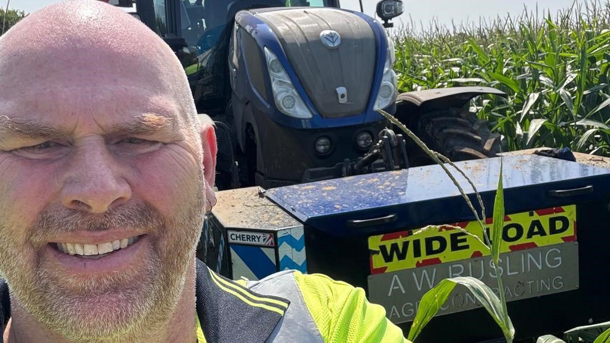 A selfie-style image of a man smiling into the camera while standing in front of a tractor in a field.