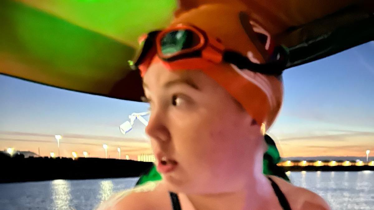 A teenage girl looks over her right shoulder while wearing an orange swimming cap and orange goggles as the sunsets behind her. She is stood on a boat as it leaves a port.