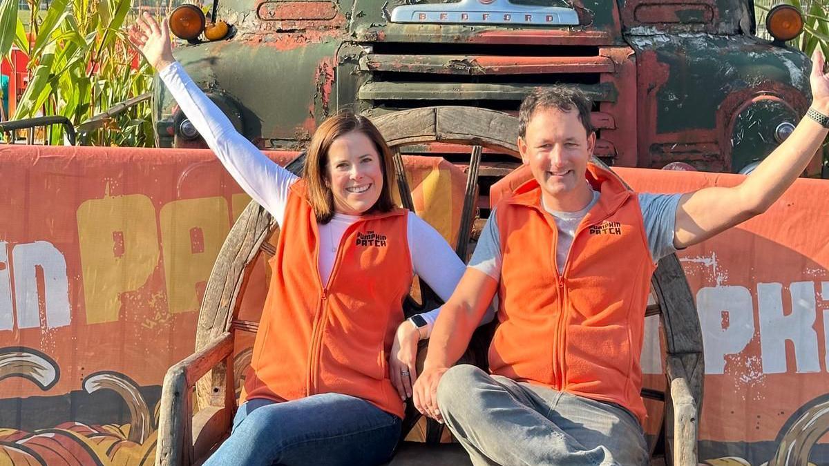 Emily French and Guy sitting down on chairs. They are both smiling with one hand up on each side. They are both wearing orange fleeces with "Pumpkin Patch" written on their lapels. 
