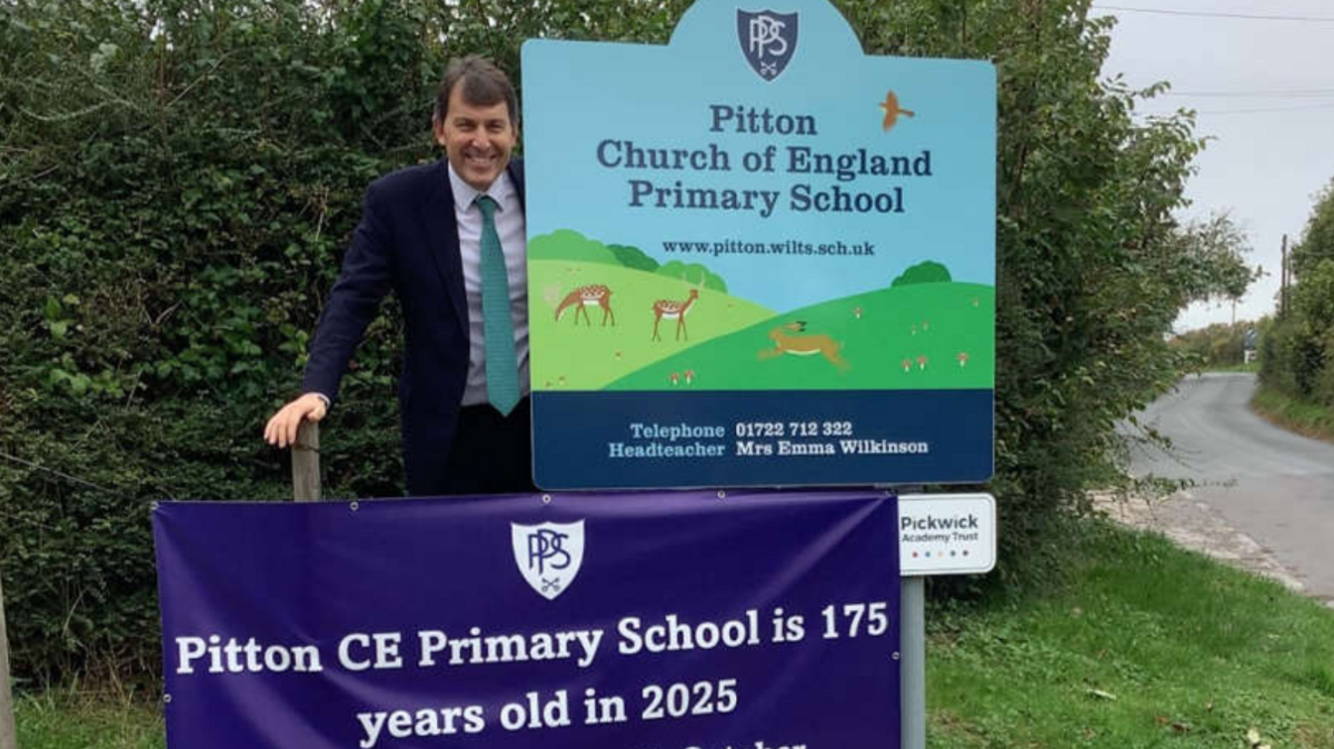 A man in a suit and green tie standing next to a sign which reads 'Pitton Church of England Primary School' with a picture of a hill and deer.