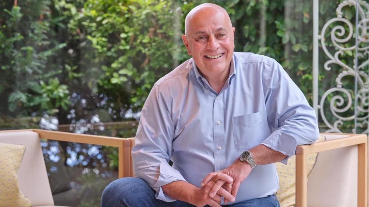 Andreas Antona, owner of Simpsons, sits in a chair at a coffee table, with a window behind him and a few of a garden with trees. He is wearing a light blue shirt, open at the neck, and jeans. He has his hands loosely clasped together and he is smiling.