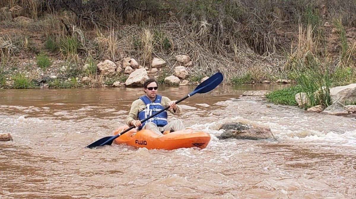 Mr Perrett is in an orange kayak in a river with greenery on the banks. He has dark hair and is wearing sunglasses. He has a blue life jacket on and is holding a blue oar.