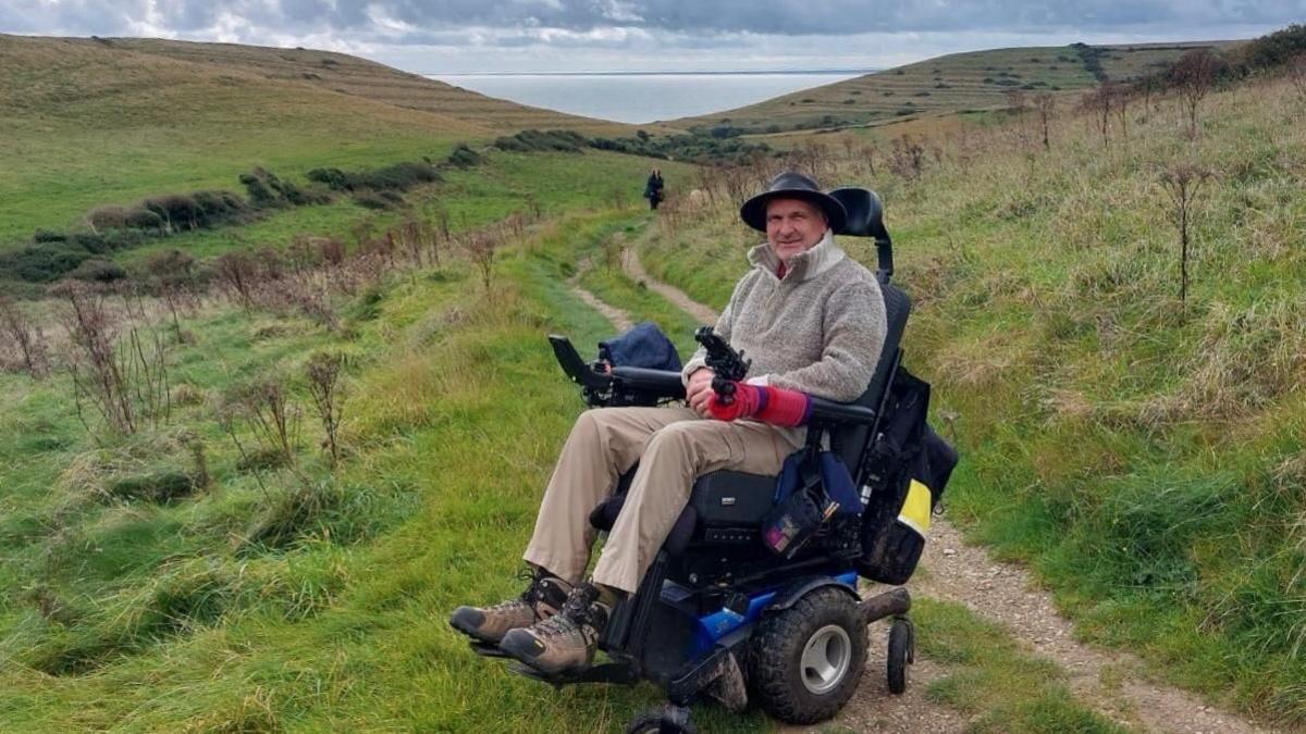 A man in a powered wheelchair parked to one side of a track running down through a field towards the sea. He is wearing a hat and looking towards the camera