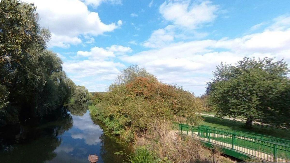 A river reflecting clouds is visible to the left, flanked by trees and hedges. A path edged with green railings is on the right with a tree in the background.