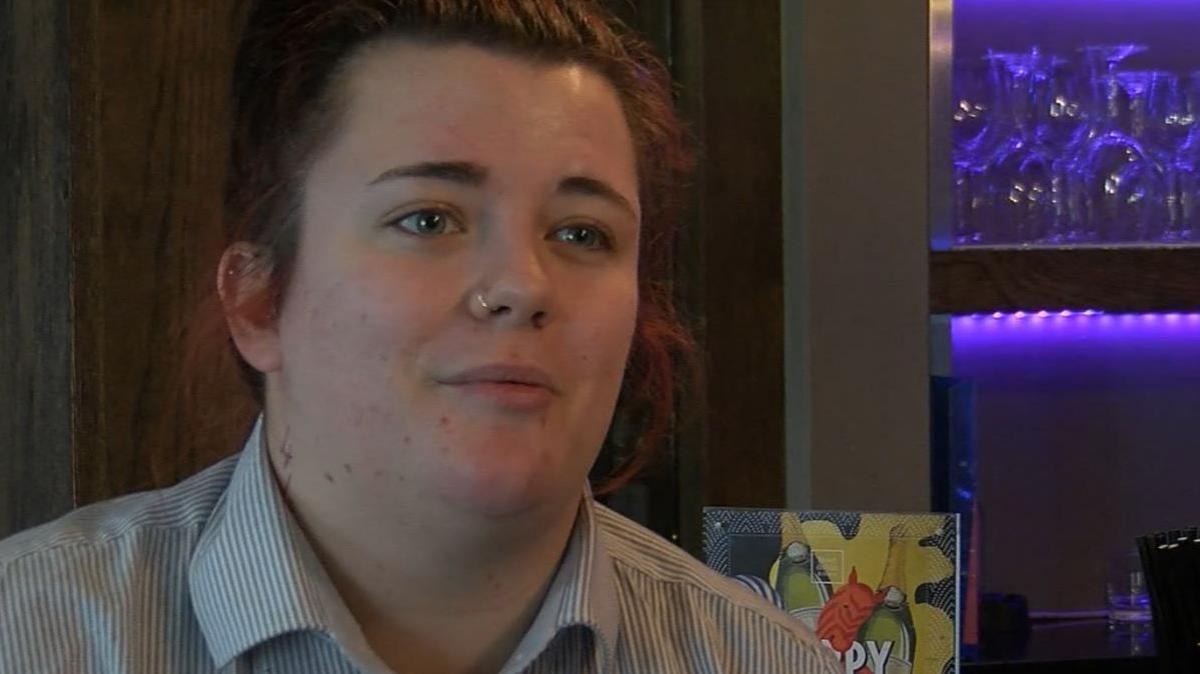 A 23 year old woman in her late teens looks into the camera. She has tied back red hair, a nose ring and is wearing a smart striped shirt. The background is wall of a pub where she works, with patterned wallpaper and wine glasses on a shelf behind her. 