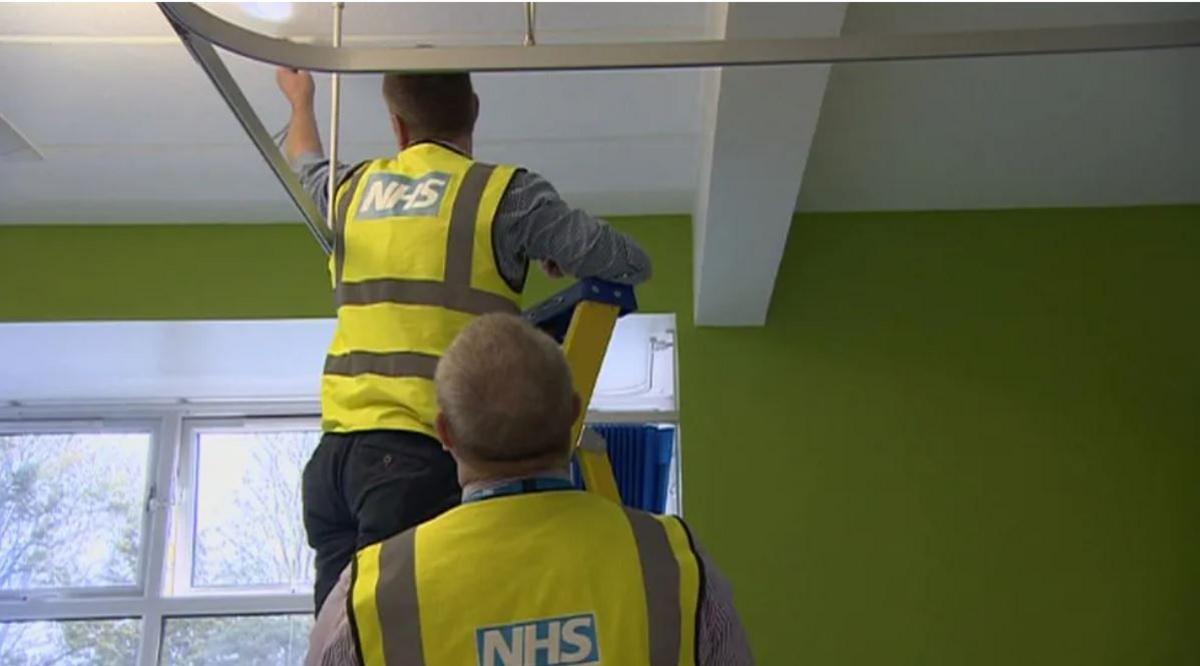 Two men in high-ves vests with NHS logos on the back in a green room, one of the men is on a ladder with his arm outstretched to the ceiling