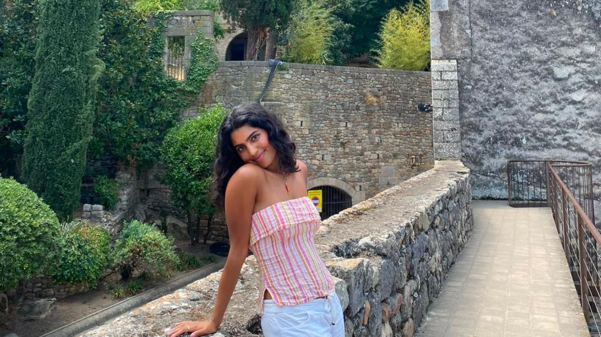 Natalina has shoulder length black hair and wears a pink strapless top with stripes. She wears an orange necklace and white trousers and is leaning against a stone wall