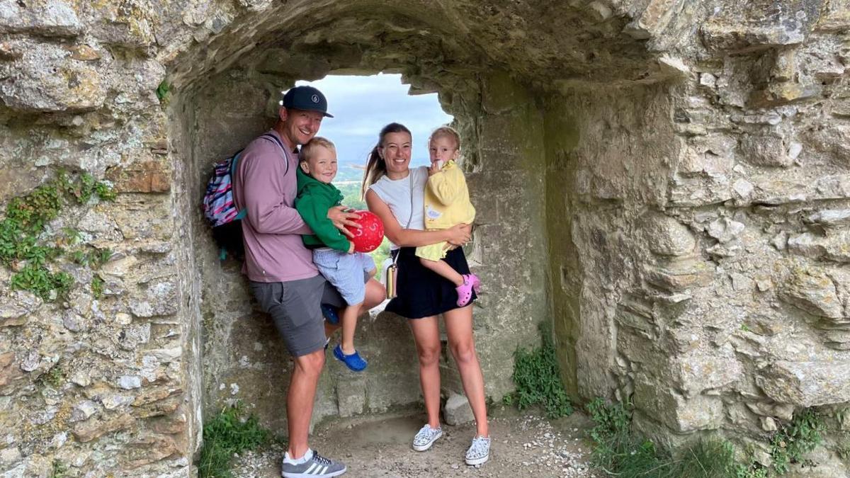 A family of a mother, father and two children smiling for the camera at one of the castle's stone walls. Both parents are holding a child. The father is holding a red ball as well.