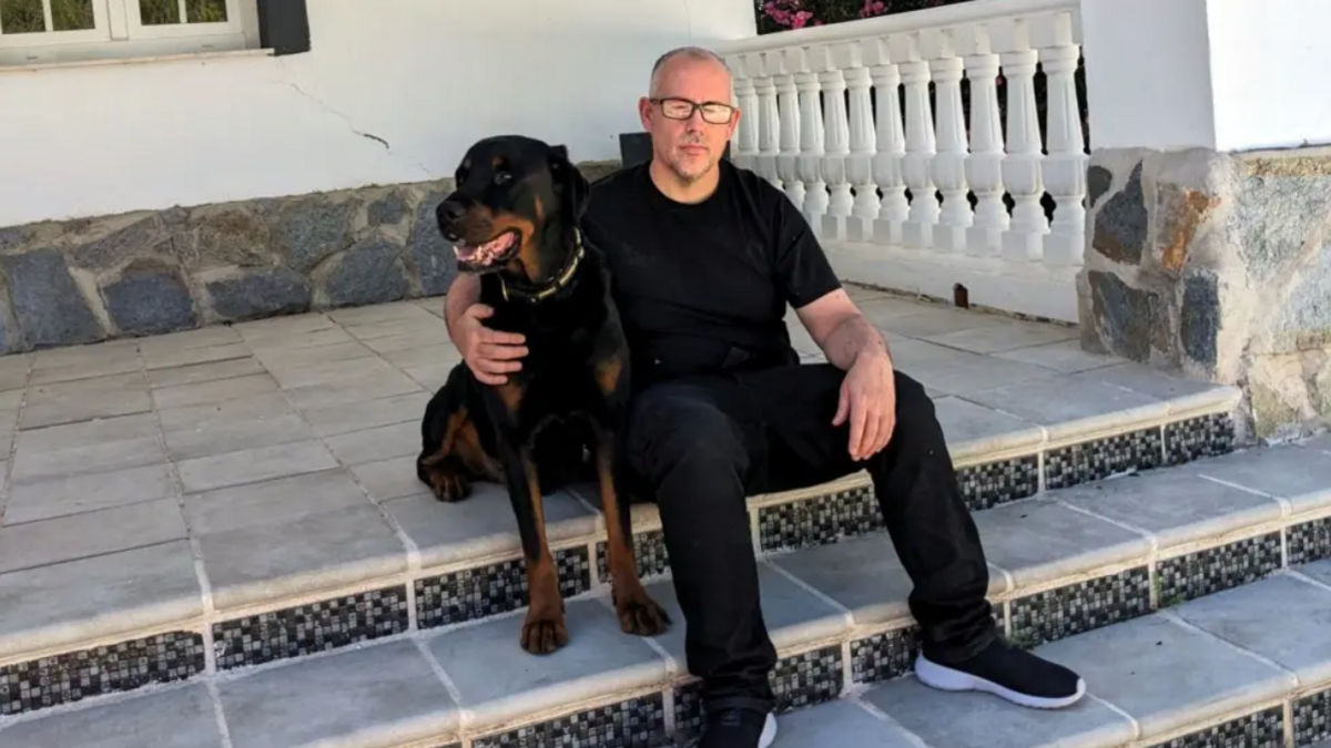 Ryan is sitting on some steps outside a building with his right arm around his dog that's also sitting on the steps. He is dressed in a black t-shirt, black trousers and black trainers, with grey cropped hair and black glasses