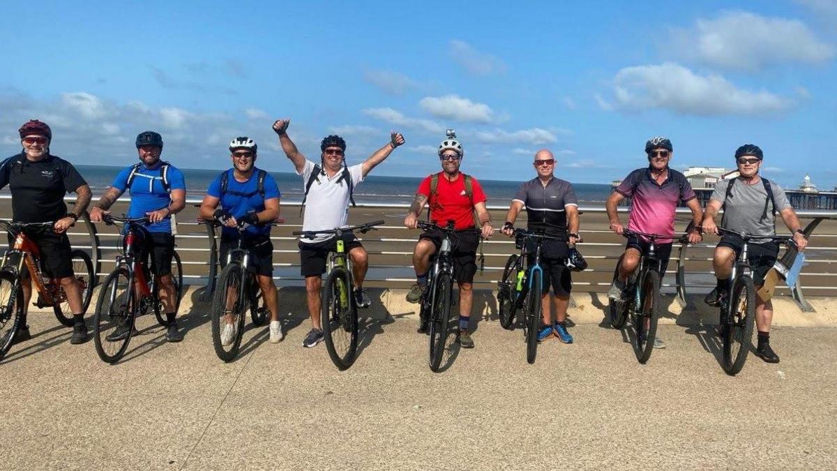 Eight men in cycling gear line up along a seafront while straddling their bikes on a sunny day. They are smiling, with one of the line-up holding their hands up in the air.