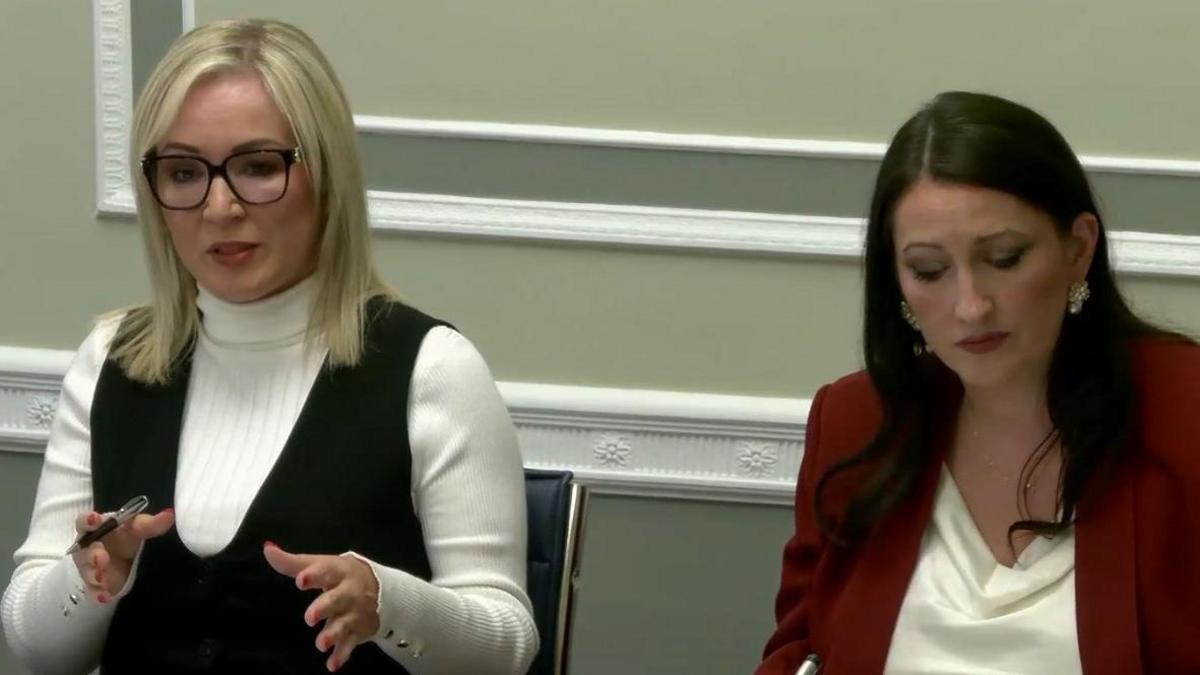 Michelle O'Neill and Emma Little-Pengelly appearing before the Executive Office committee at Stormont on Wednesday. O'Neill has straight blonde hair and is wearing a white turtleneck jumper under a black dress or tanktop. Little-Pengelly has long, dark hair and is wearing a red jacket and a white top.