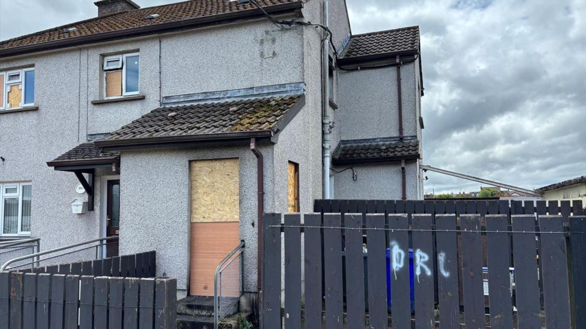 A grey end-terrace house with a boarded up door and windows. There is a grey fence in front of the house and a cloudy sky above. 