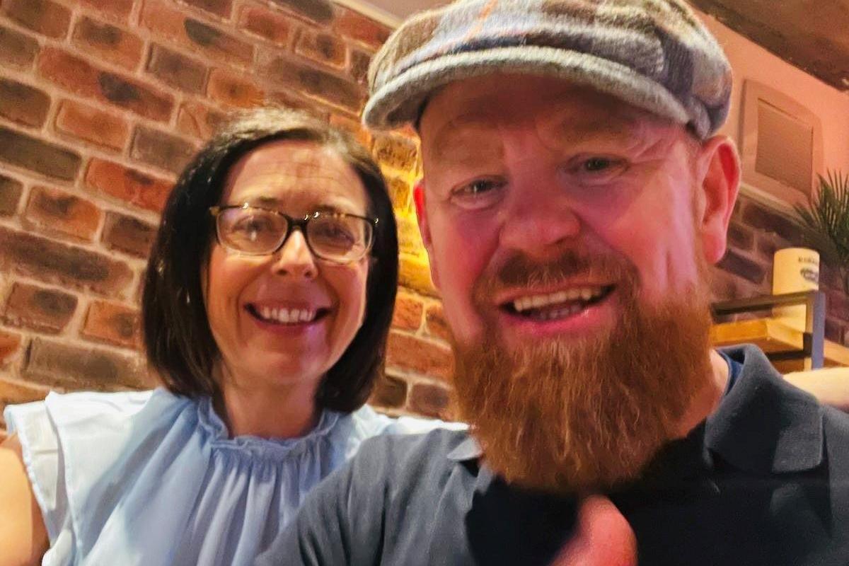 Man with ginger beard, wearing a bunnet, wearing a dark top and holding his thumb up, smiling, with a smiling woman with side-parted dark hair and glasses, wearing a short-sleeved light blue blouse, with her hand round his upper arm.