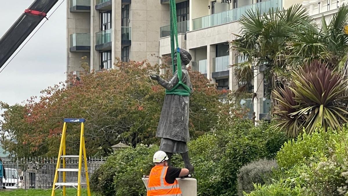 An engineer wearing a white helmet and orange high-vis jacket inspects a bronze statue of Nancy Astor before a small crane with a green harness lifts the structure from its plinth. The green harness is wrapped around the centre of the statue.
