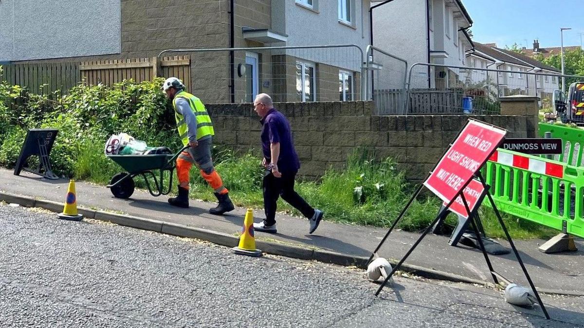 A man in hi vis with a wheelbarrow with household items followed by another man