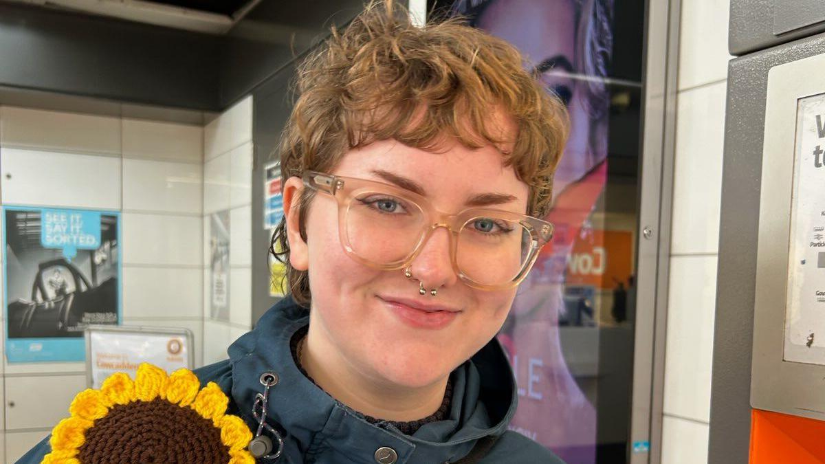 Elise Prentice smiles at the camera, wearing large fashionable glasses, a navy jacket with a knitted sunflower pinned on the right hand side. Behind, we can see a Glasgow underground station with safety posters on the wall.