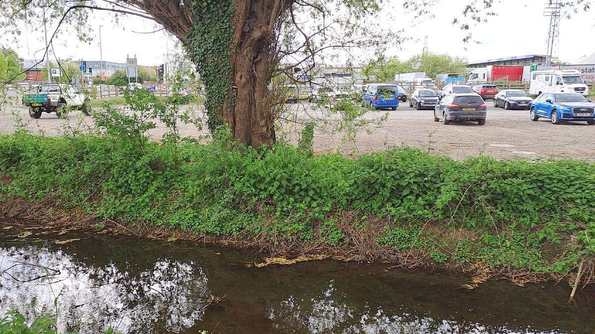A shallow brook runs adjacent to a small car park with Hereford FC's stadium in the background.