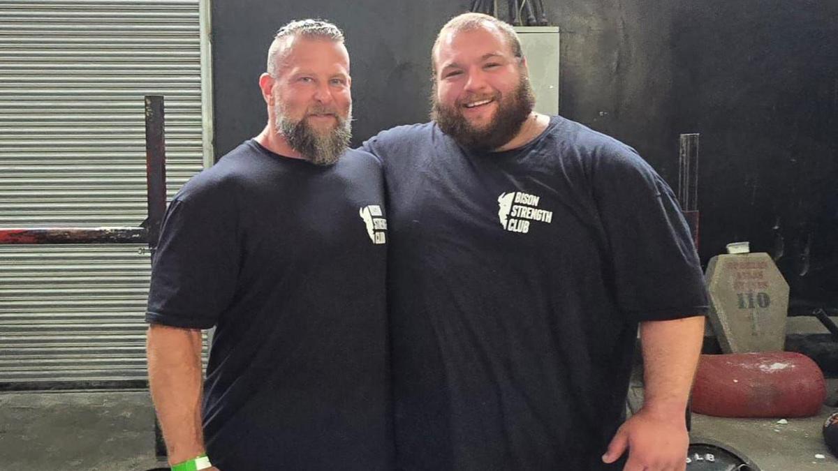 Two men stand together with their arms around each other. Both are smiling. They are wearing dark T-shirts and are in a gym.
